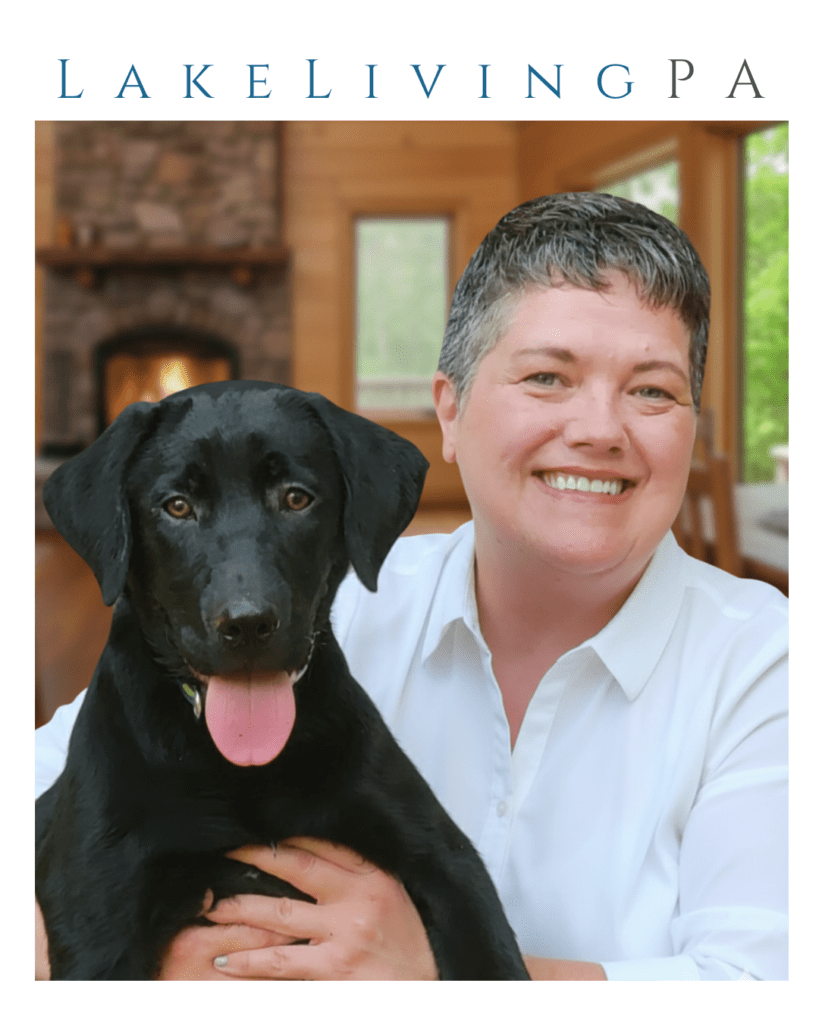 Realtor Karen Rice in a white shirt smile sand holds a black Labrador retriever in a cozy, well-lit room with a fireplace, highlighting a warm, friendly atmosphere.